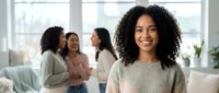 Happy young African American woman smiling in modern office with diverse female colleagues collaborating in background during business meeting.