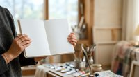 Artist holding open blank sketchbook in studio workspace with paintbrushes, art supplies, and natural window light for creative projects and design work.