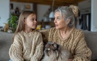 Grandmother and granddaughter in matching knit sweaters sharing a tender moment with a fluffy cat on cozy sofa at home.