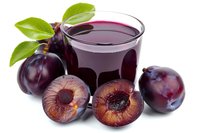 Fresh plum juice in glass with whole and halved plums featuring green leaves, isolated on white background for beverage, food and drink design projects.