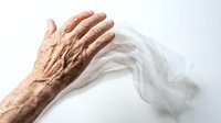 Elderly hand with wrinkled skin touching a delicate translucent fabric on a white background, symbolizing fragility and aging.