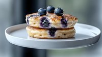 Fresh blueberry pancakes stacked on white ceramic plate, topped with fresh berries. Golden brown surface with visible fruit pieces, soft fluffy texture.