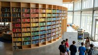 Modern public library interior with curved wooden bookshelves filled with colorful books and students studying below in bright natural light space.
