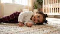 Happy mixed-race toddler boy with curly hair lying on soft carpet in bright nursery room, wearing plaid pajamas and smiling at camera in cozy home setting.
