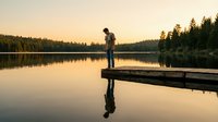 Young man standing on wooden dock at serene lake during golden hour sunset, peaceful water reflection, forest background for relaxation concepts.