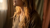 Young blonde Caucasian woman in cozy sweater looking thoughtfully out window with warm golden sunlight streaming across her face and long wavy hair.