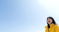 Young woman with dark hair and East Asian ethnicity wearing bright yellow jacket, smiling outdoors against clear blue sky. Suitable for lifestyle, happiness, and wellness content.