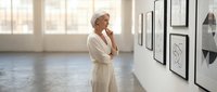 Thoughtful senior Caucasian woman in white blazer contemplating framed artwork in modern gallery space with natural lighting for business presentations.