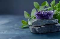 Raw amethyst crystal cluster on stacked dark stones with fresh green leaves against blue textured background, creating zen-like spa atmosphere.