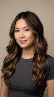 Young Asian businesswoman with wavy brown hair and highlights wearing dark shirt smiling confidently against neutral beige background for professional headshot.