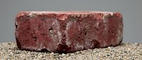 Red clay brick with weathered texture and rough edges sitting on gravel surface, construction material for building and masonry projects.