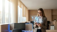 Young Caucasian businesswoman in glasses using smartphone with holographic cybersecurity interface in modern office, digital data protection concept.