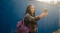 Young African American woman with dreadlocks taking selfie with smartphone outdoors, wearing casual jacket and purple backpack in bright sunlight.