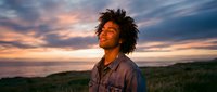 Young African American man with dreadlocks looking up at colorful sunset sky outdoors, expressing joy and optimism in natural landscape setting.
