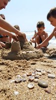 Family building sandcastle together at beach with ocean waves in background, children and adults enjoying summer vacation outdoor activity.