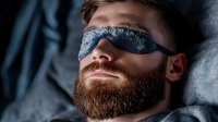 Caucasian bearded man wearing sleep mask resting on pillow in dark room. Peaceful facial expression shows deep relaxation during nighttime rest period.