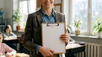 Professional Caucasian female teacher holding clipboard in bright classroom with students at desks, educational environment for academic presentations.