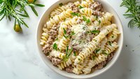 Creamy fusilli pasta with ground meat sauce, garnished with fresh rosemary and parsley in white ceramic bowl on light background. Perfect for recipe content.