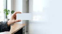 Professional holding blank white card in hand against modern office background with natural light and green plant, suitable for business branding and corporate messaging.