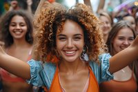 Young woman with curly hair and bright smile enjoying outdoor festival celebration, wearing orange top and blue blouse, expressing joy and happiness.