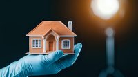 Hand in medical glove holding miniature orange house model against dark background with glowing light, representing real estate healthcare or property protection.