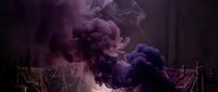 Dramatic purple and violet smoke clouds billowing in a dark, moody studio setting with candles and draped fabric in the background.