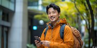 Asian man in orange jacket using smartphone while walking city street, wearing backpack, casual urban lifestyle, smiling at camera in autumn setting.