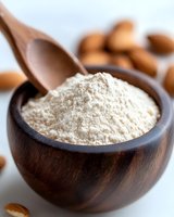 Fresh almond flour in wooden bowl with raw almonds scattered around, macro food photography on light background for healthy baking and cooking concepts.