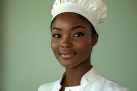 Young African American female chef in white uniform and traditional toque smiling confidently against green background, professional culinary portrait.