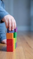 Child hand placing colorful wooden blocks in tower formation on wooden floor demonstrating early learning development and fine motor skills practice.