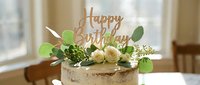 Rustic naked birthday cake decorated with white roses, eucalyptus greenery, and a wooden Happy Birthday topper on a bright table.