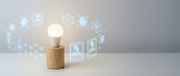 Glowing light bulb on wooden base with floating digital icons representing innovation, technology and creative business solutions for modern workspace.