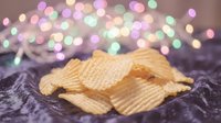 Golden crispy potato chips scattered on soft purple fabric with colorful bokeh lights creating festive party atmosphere for snack food advertising.