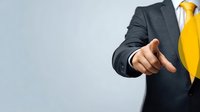 Businessman in dark suit with bright yellow tie pointing finger forward against neutral background, conveying leadership and decision-making in corporate environment.