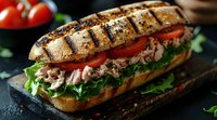 Grilled tuna sandwich with fresh lettuce, tomato slices, and herbs on toasted artisan bread, served on dark stone surface with fresh vegetables in background.