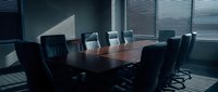 Modern corporate boardroom with dark wooden conference table and black leather chairs illuminated by natural light through venetian blinds.