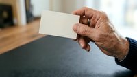 Senior man with dark skin tone holding blank white business card in hand at wooden desk, suitable for corporate identity, networking, and professional branding concepts.