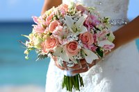 Romantic beach wedding bouquet featuring pink roses, white lilies, and delicate greenery held by bride in white dress against turquoise ocean background.