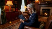 Middle-aged woman with gray hair and glasses wearing a dark blazer, holding a tablet while seated in an upscale library study room with fireplace, wooden furniture, and warm lighting for business, technology, and professional concepts.