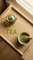 Matcha green tea ceremony flatlay on wooden tray with glass teapot, bamboo whisk, ceramic bowl and TEA lettering made of matcha powder.