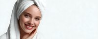 Young woman with bright smile wearing white towel on head after shower, touching face with hand against clean background. Perfect for beauty and skincare concepts.