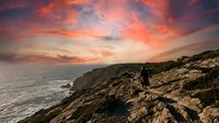 Woman in casual attire standing on rocky coastal cliff at sunset, gazing toward ocean horizon with dramatic orange and pink sky, golden hour landscape photography for travel and lifestyle use.