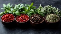 Fresh aromatic herbs and colorful peppercorns in clay bowls on dark background, pink, black, and green pepper varieties with basil leaves for cooking.