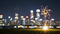 Sparkler burning bright on grass with blurred city skyline at night, celebration and festive atmosphere for holidays and special occasions.
