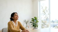 Woman with dark hair and South Asian ethnicity, wearing a mustard yellow cardigan, sitting indoors near a window with potted plants visible. Office or home workspace setting with natural light. Professional, content, and relaxed atmosphere.