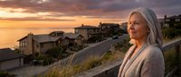 Mature woman with long white hair and light skin tone, wearing a cardigan, standing on a coastal hillside road at sunset overlooking luxury homes and ocean views during golden hour.