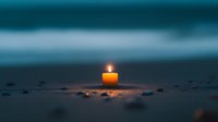 Solitary glowing candle on dark beach sand against moody teal ocean horizon, creating peaceful ambiance for meditation or remembrance ceremonies.