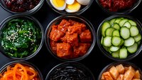 Korean side dishes in black bowls featuring kimchi, seaweed, cucumber, marinated vegetables, boiled eggs and fermented specialties arranged in traditional banchan style.