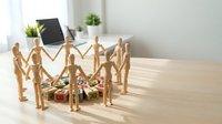 Wooden figurines standing in circle around coins and dollar bills on desk with laptop and office supplies in background, representing teamwork, financial planning, and business collaboration.