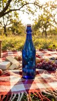 Blue glass bottle on checkered blanket with cheese and grapes for outdoor picnic in sunny park setting with warm golden light filtering through trees.